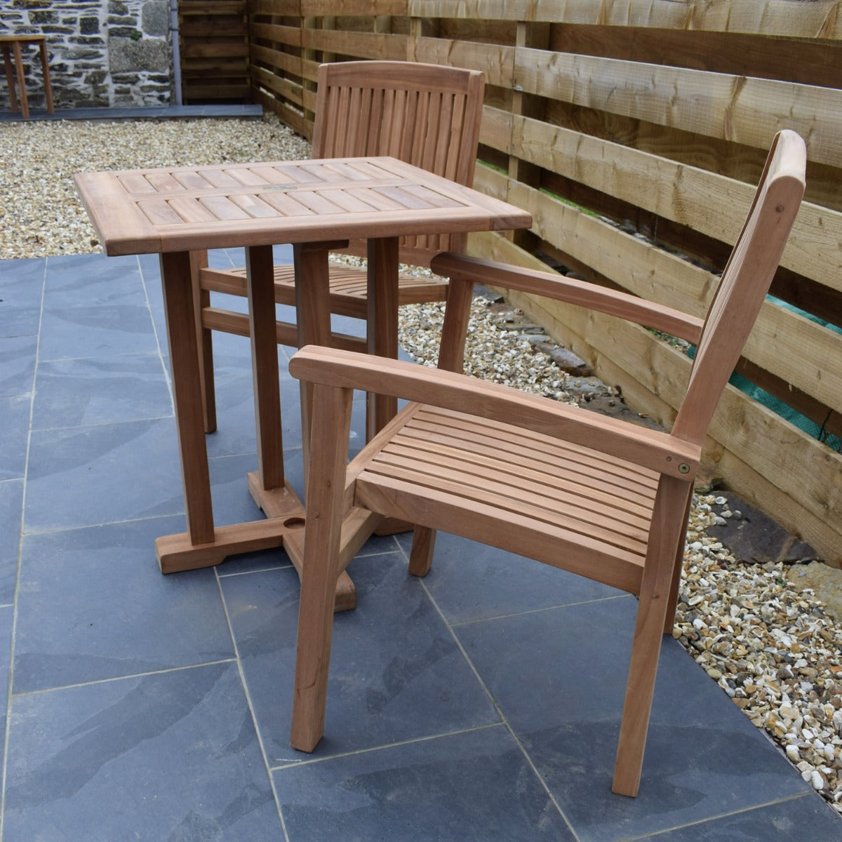 side view of 70cm teak square pedestal garden dining table with 2 Henley style stacking garden armchairs